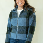 A woman modeling a blue and black buffalo plaid zip-up fleece jacket, paired with dark blue jeans and tan boots, standing against a plain off-white studio background.