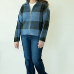 A woman modeling a blue and black buffalo plaid zip-up fleece jacket, paired with dark blue jeans and tan boots, against a plain white studio background.