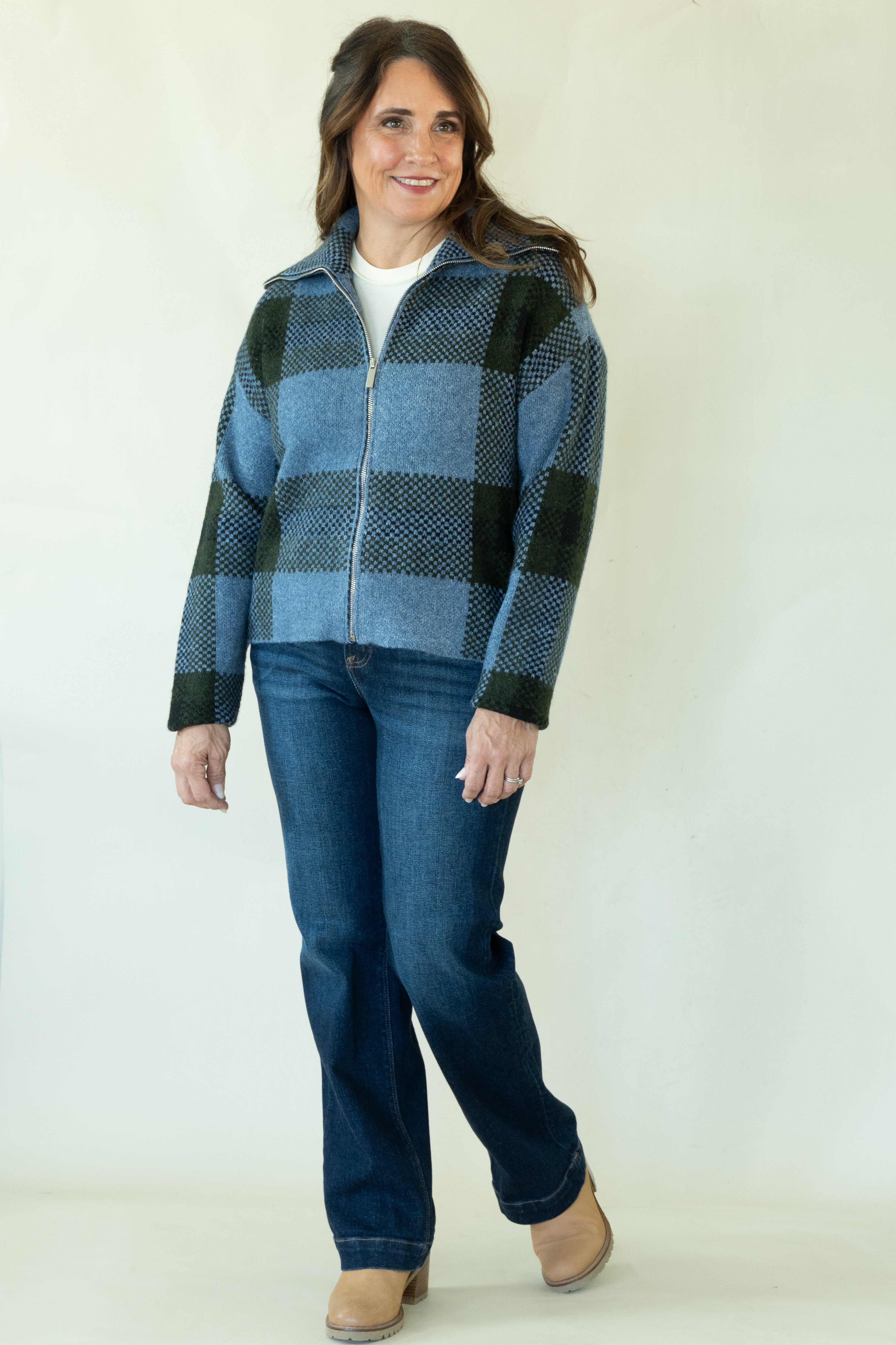 A woman modeling a blue and black buffalo plaid zip-up fleece jacket, paired with dark blue jeans and tan boots, against a plain white studio background.