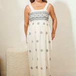 Woman wearing a white dress with black patterns standing against a plain wall.