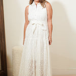 Woman wearing a white sleeveless dress with a belt, standing in a room with a mirror and textured wall.