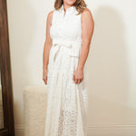 Woman wearing a white sleeveless dress with a belt, standing against a plain wall.