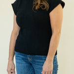 A woman wearing a black short-sleeve mock-neck knit sweater and blue jeans, standing against a plain light beige background.