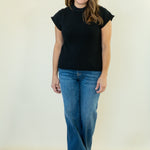 A woman modeling a black short-sleeve mock neck top and blue jeans against a plain off-white studio background.