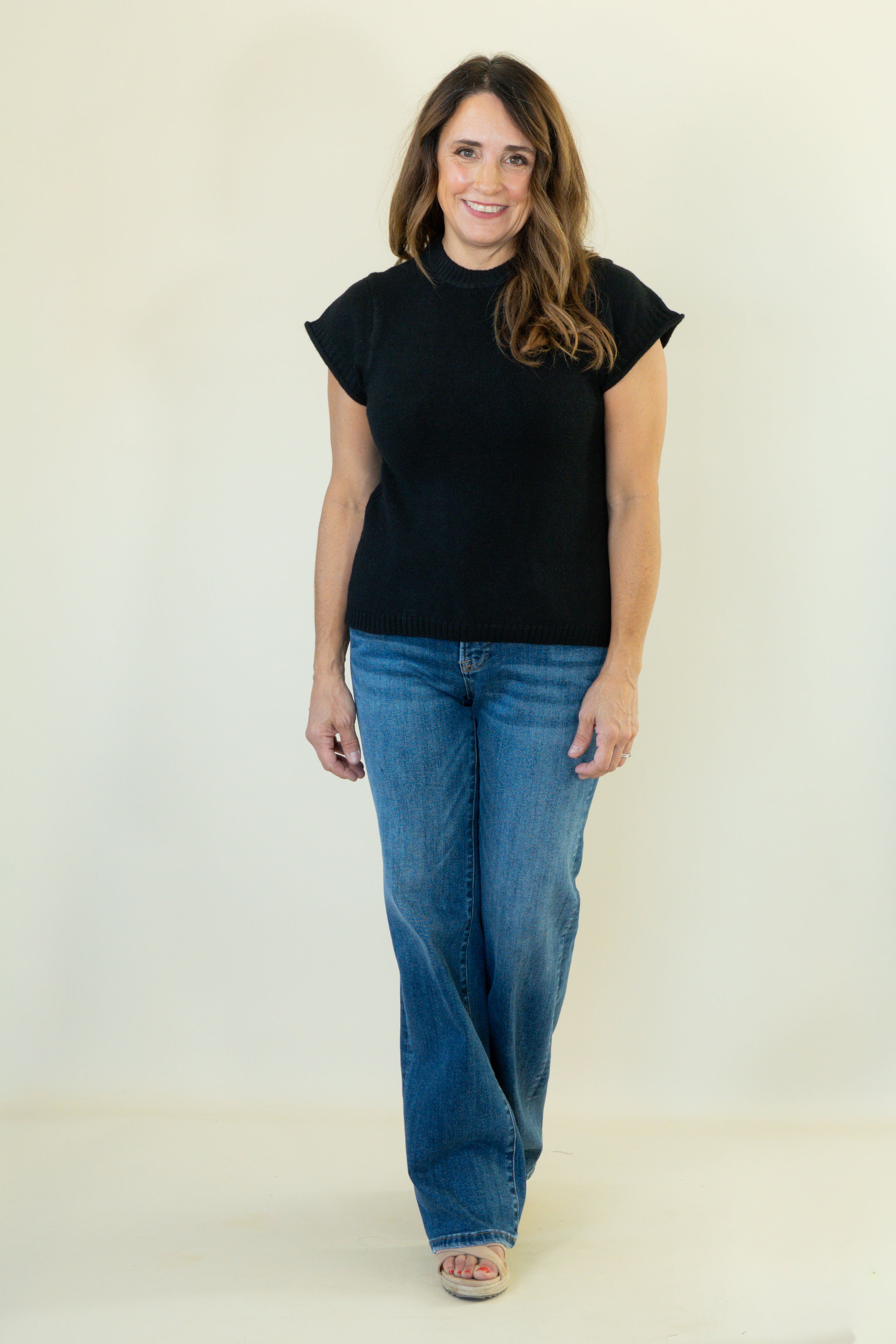 A woman modeling a black short-sleeve mock neck top and blue jeans against a plain off-white studio background.