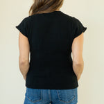 A woman viewed from behind wearing a black short-sleeve knit top and blue jeans against a plain light beige background.