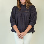 A woman wearing a dark navy eyelet blouse with puff sleeves and white pants, standing against a plain light beige background.