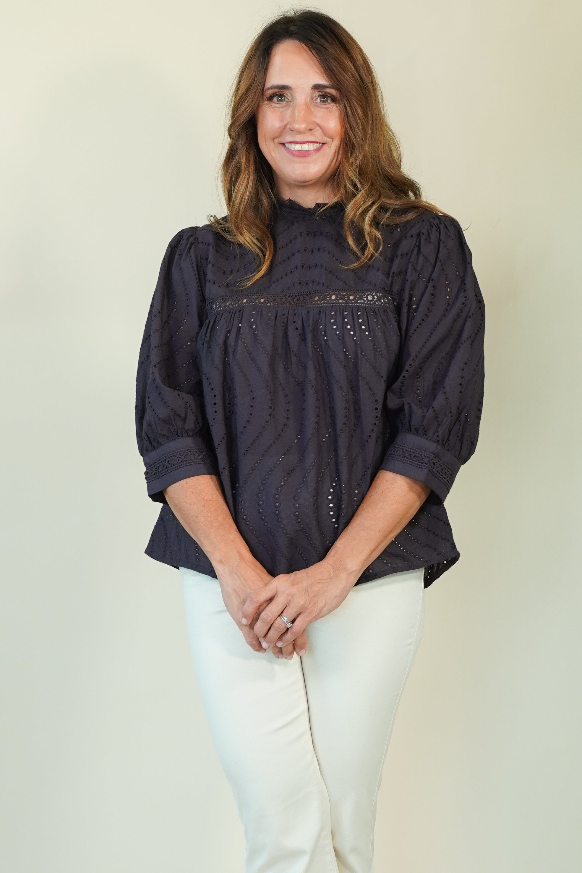 A woman wearing a dark navy eyelet blouse with puff sleeves and white pants, standing against a plain light beige background.
