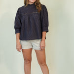 A woman modeling a dark navy blue textured long-sleeve blouse with eyelet details on the chest, paired with light beige shorts and tan sandals, standing against a plain light beige studio background.