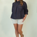 A woman modeling a dark navy blue textured long-sleeve blouse with white shorts and tan slide sandals, standing against a plain light beige studio background.