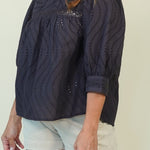 A woman wearing a dark navy eyelet blouse with textured vertical eyelet stripes and a high mock neck, paired with light beige shorts, against a plain beige background.