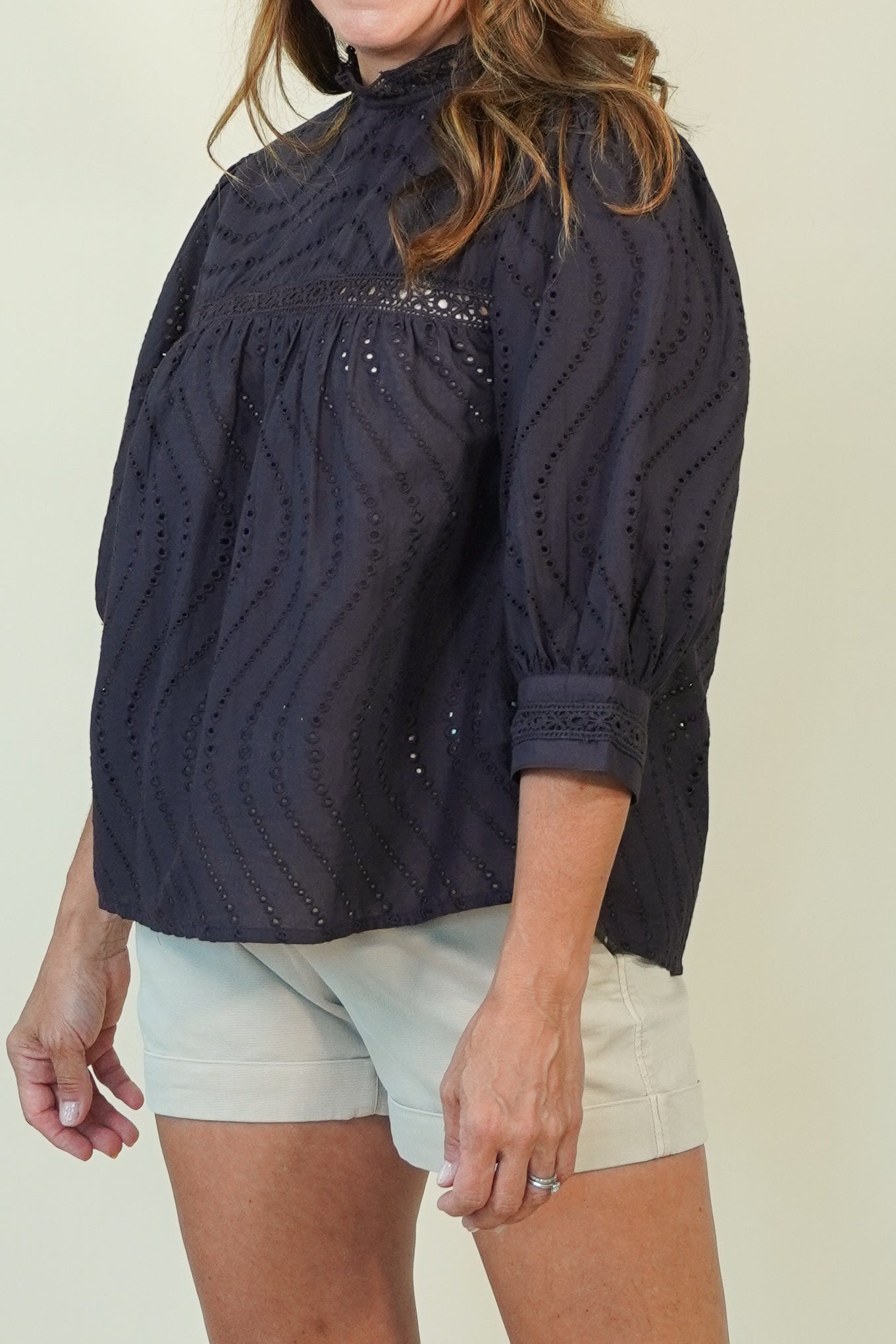 A woman wearing a dark navy eyelet blouse with textured vertical eyelet stripes and a high mock neck, paired with light beige shorts, against a plain beige background.