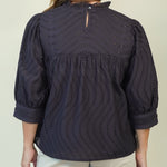 A woman shown from the back wearing a dark navy eyelet blouse with three-quarter sleeves and light beige shorts, set against a plain light beige background.