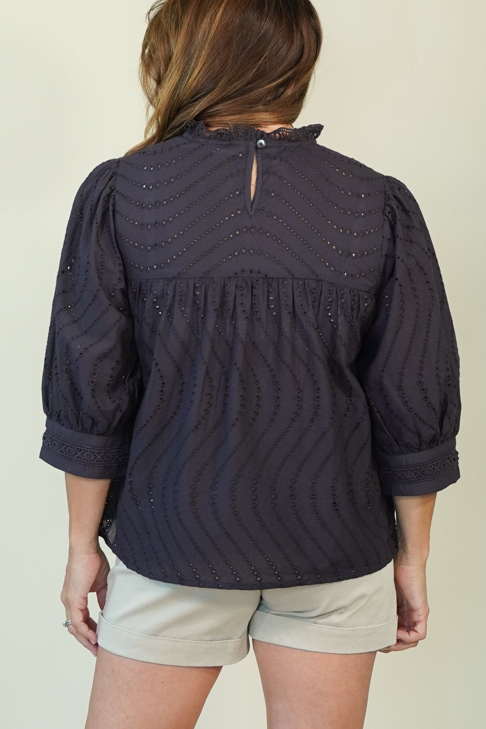 A woman shown from the back wearing a dark navy eyelet blouse with three-quarter sleeves and light beige shorts, set against a plain light beige background.