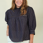 A woman wearing a dark navy eyelet blouse with three-quarter sleeves and white pants, standing against a plain light beige background.