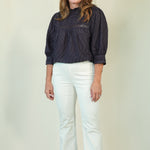 A woman modeling a dark navy blue textured blouse with three-quarter sleeves and white pants, standing against a plain light beige studio background.