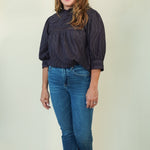 A woman modeling a dark navy textured blouse with puff sleeves and blue jeans, standing against a plain light beige studio background.