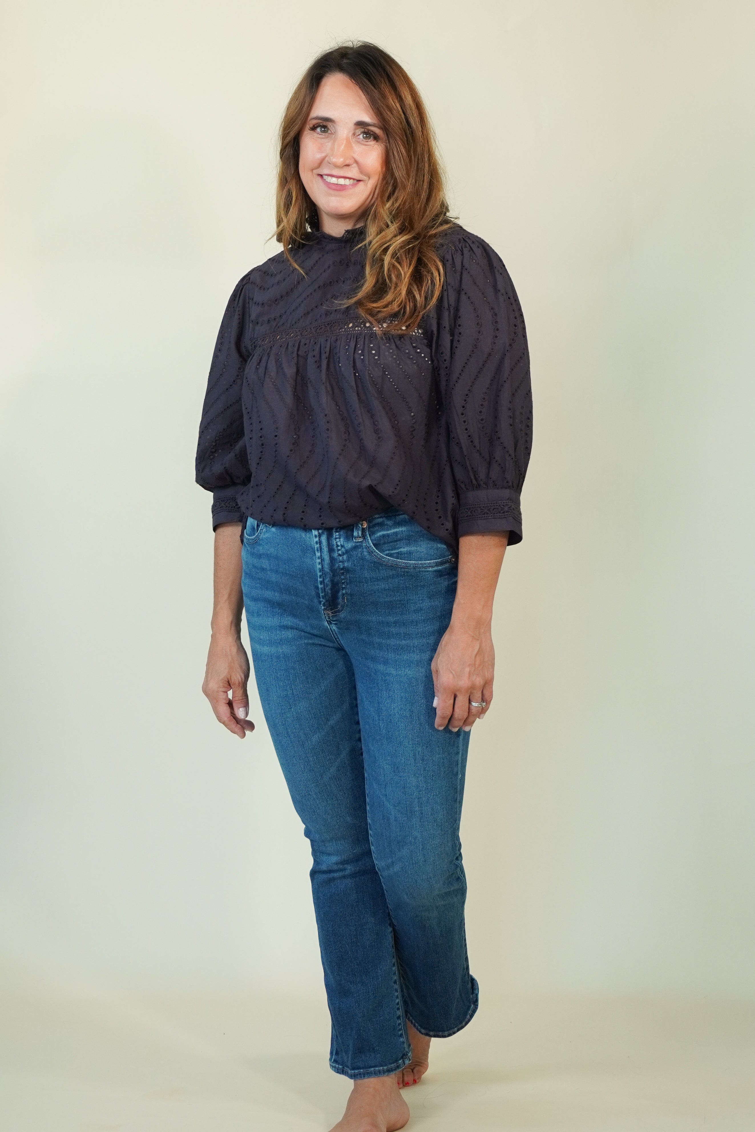 A woman modeling a dark navy textured blouse with puff sleeves and blue jeans, standing against a plain light beige studio background.