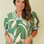 A woman wearing a green and cream abstract patterned blouse with short puff sleeves and a V-neck, paired with light beige pants, standing against a plain cream-colored wall.