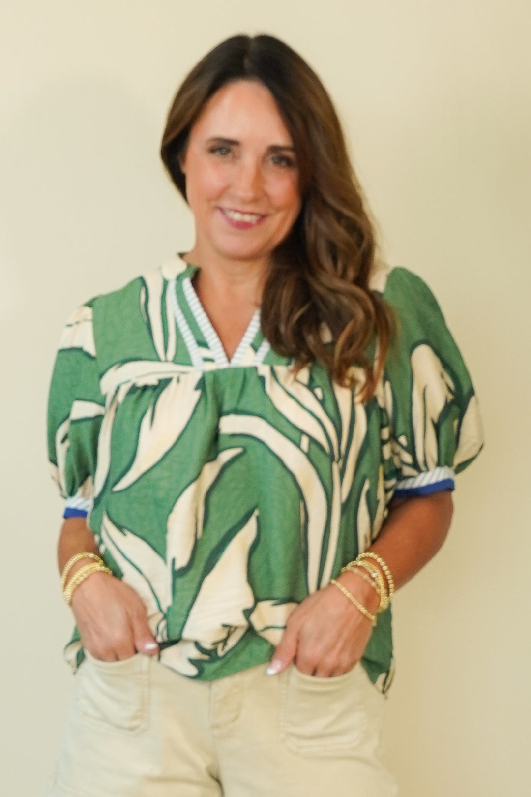 A woman wearing a green and cream abstract patterned blouse with short puff sleeves and a V-neck, paired with light beige pants, standing against a plain cream-colored wall.