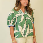 A woman wearing a green and cream tropical leaf print short-sleeve blouse with blue trim on the cuffs and collar, paired with light beige shorts, standing against a plain beige background.