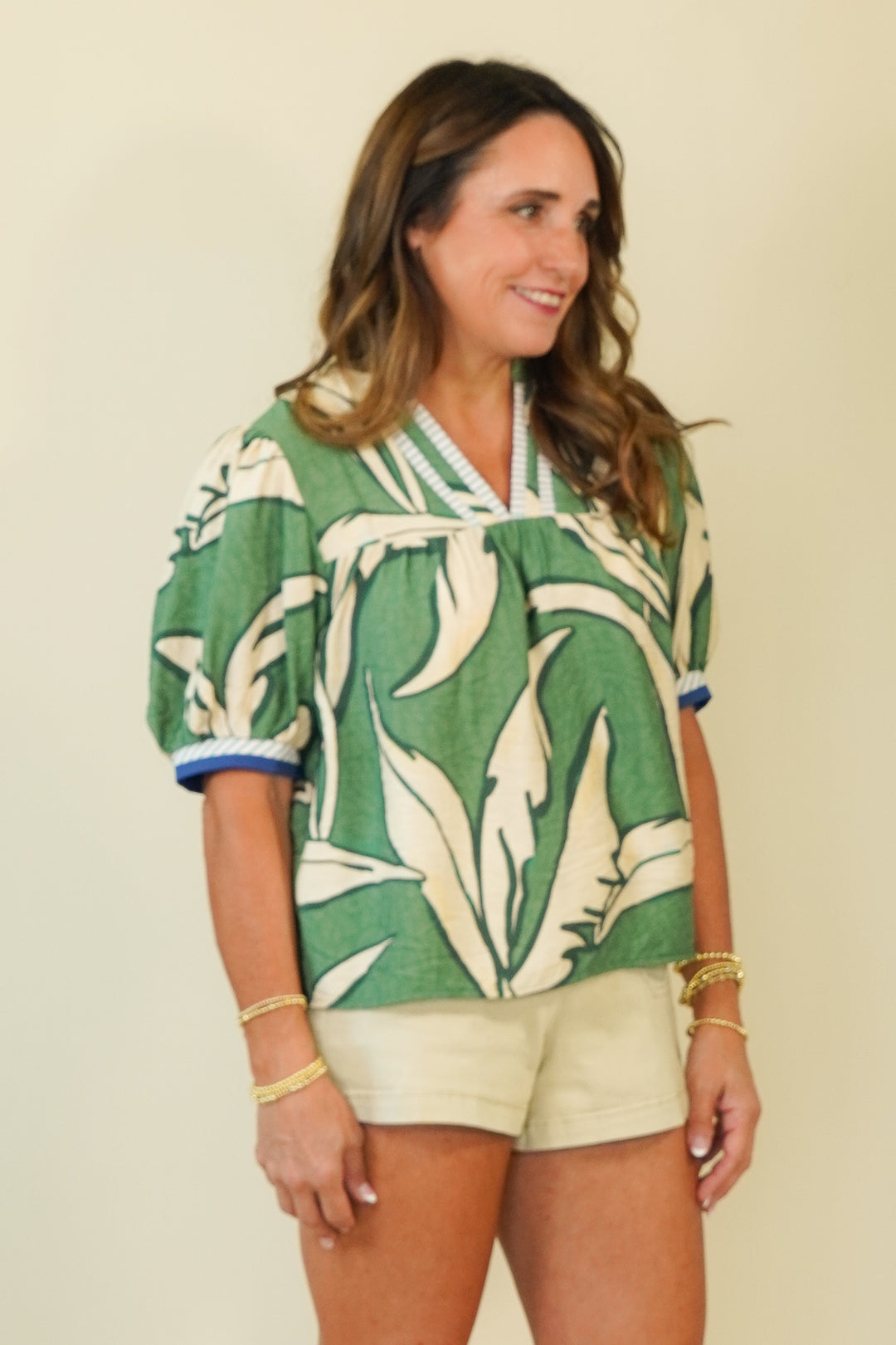 A woman wearing a green and cream tropical leaf print short-sleeve blouse with blue trim on the cuffs and collar, paired with light beige shorts, standing against a plain beige background.