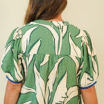 A woman viewed from the back wearing a green and white tropical leaf print short-sleeve blouse with blue trim on the sleeve cuffs, paired with light beige pants, against a plain beige background.