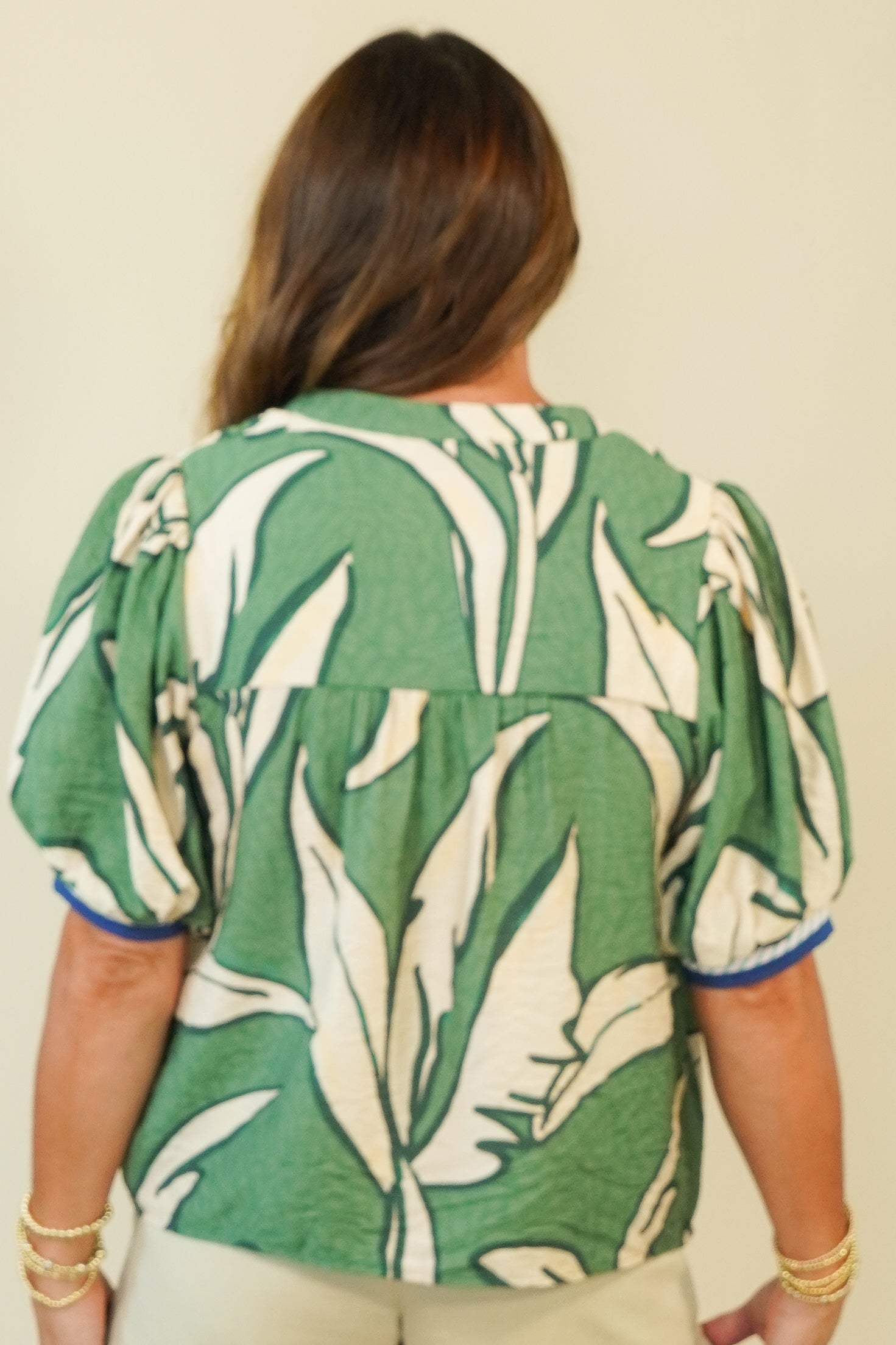 A woman viewed from the back wearing a green and white tropical leaf print short-sleeve blouse with blue trim on the sleeve cuffs, paired with light beige pants, against a plain beige background.