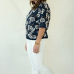 A woman modeling a navy blue short-sleeve knit top with a white floral pattern, paired with white pants and brown woven loafers, standing against a plain off-white studio background.