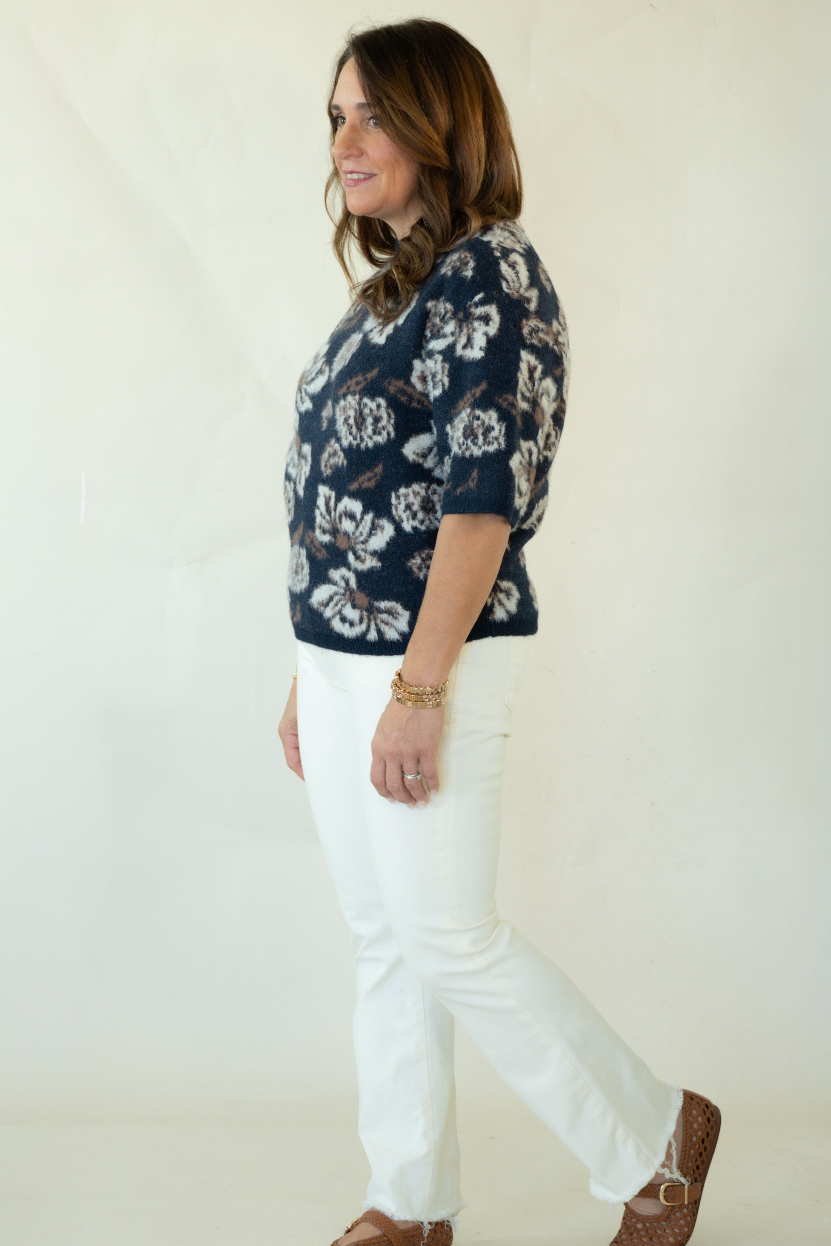 A woman modeling a navy blue short-sleeve knit top with a white floral pattern, paired with white pants and brown woven loafers, standing against a plain off-white studio background.