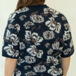 A woman viewed from behind wearing a navy blue short-sleeve button-up shirt with a white floral pattern, white pants, and brown woven sandals, standing against a plain off-white background.
