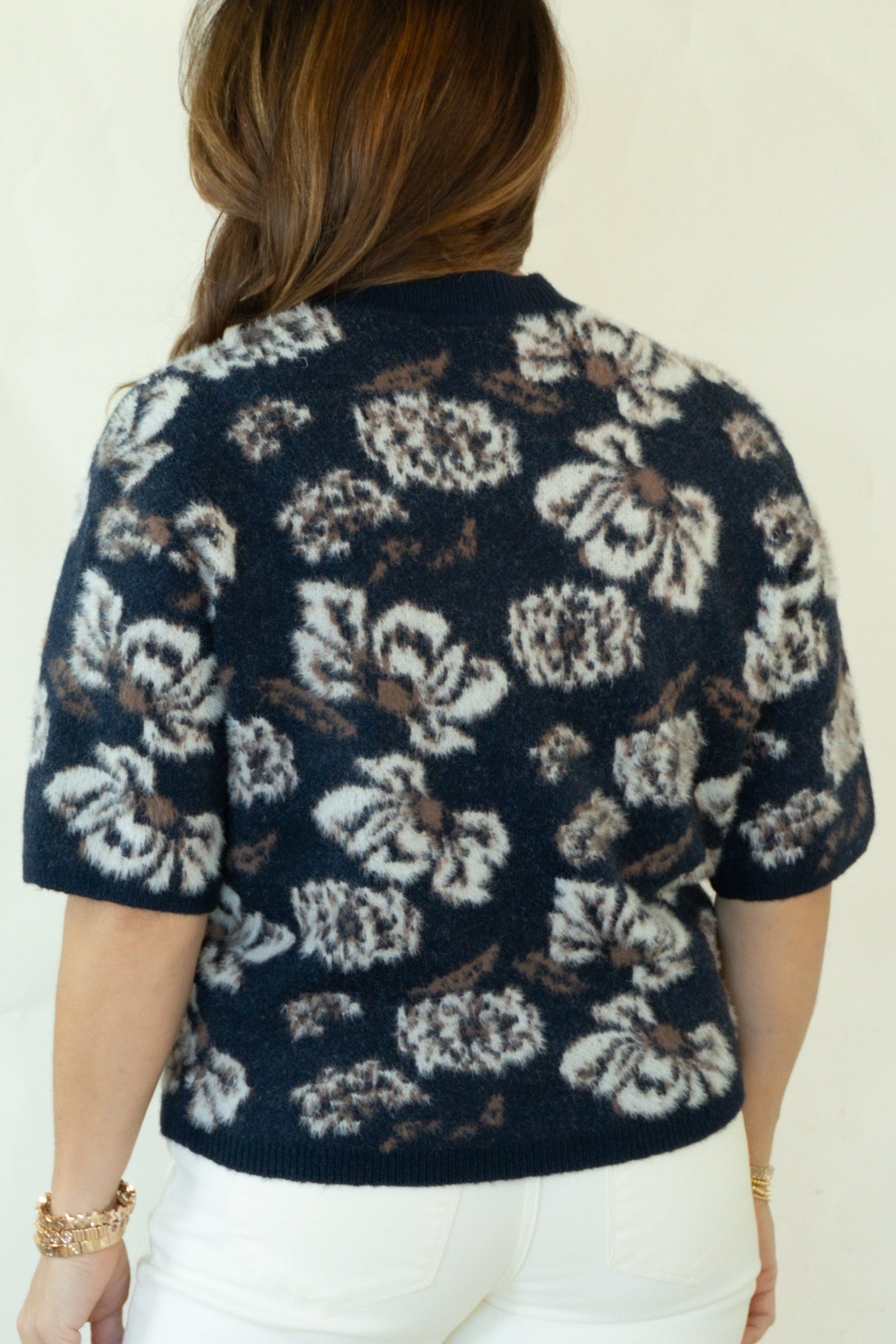 A woman viewed from behind wearing a navy blue short-sleeve button-up shirt with a white floral pattern, white pants, and brown woven sandals, standing against a plain off-white background.