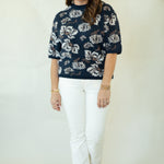 A woman modeling a navy blue short-sleeve knit sweater with a white floral pattern, paired with white pants and brown flat shoes, standing against a plain off-white studio backdrop.