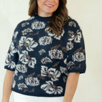 A woman modeling a navy blue short-sleeve knit top with a white floral pattern, paired with white pants and brown woven shoes, standing against a plain off-white studio background.