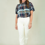 A woman modeling a short-sleeve plaid blouse with navy, red, and light blue stripes, paired with off-white pants and beige sandals, against a plain beige studio background.