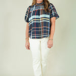 A woman modeling a short-sleeve plaid blouse with navy, red, and light blue stripes, paired with off-white pants and beige sandals, against a plain beige studio background.