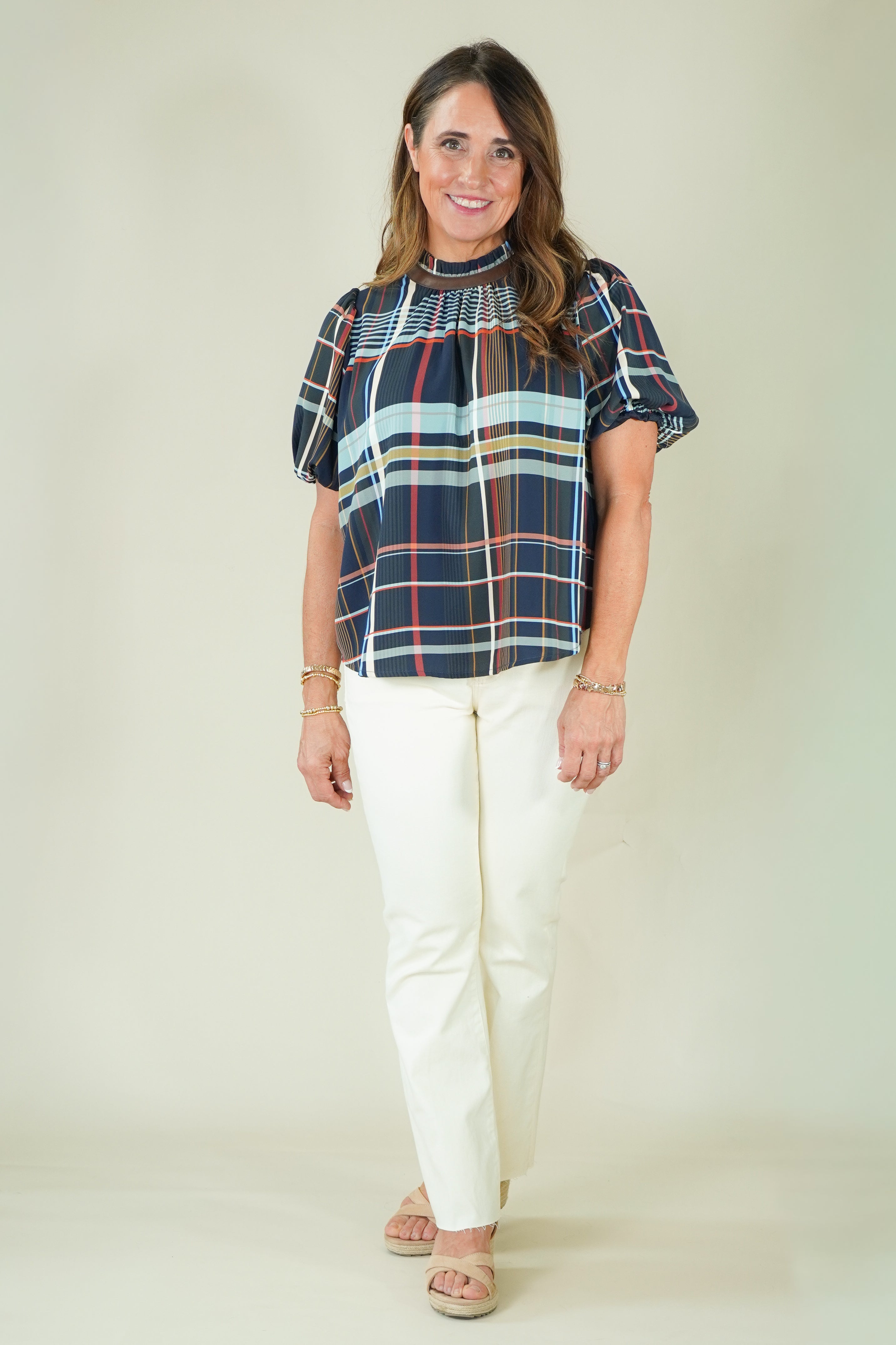 A woman modeling a short-sleeve plaid blouse with navy, red, and light blue stripes, paired with off-white pants and beige sandals, against a plain beige studio background.
