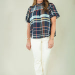 A woman modeling a short-sleeve plaid blouse with navy, red, and light blue stripes, paired with off-white pants and beige sandals, against a plain beige studio background.