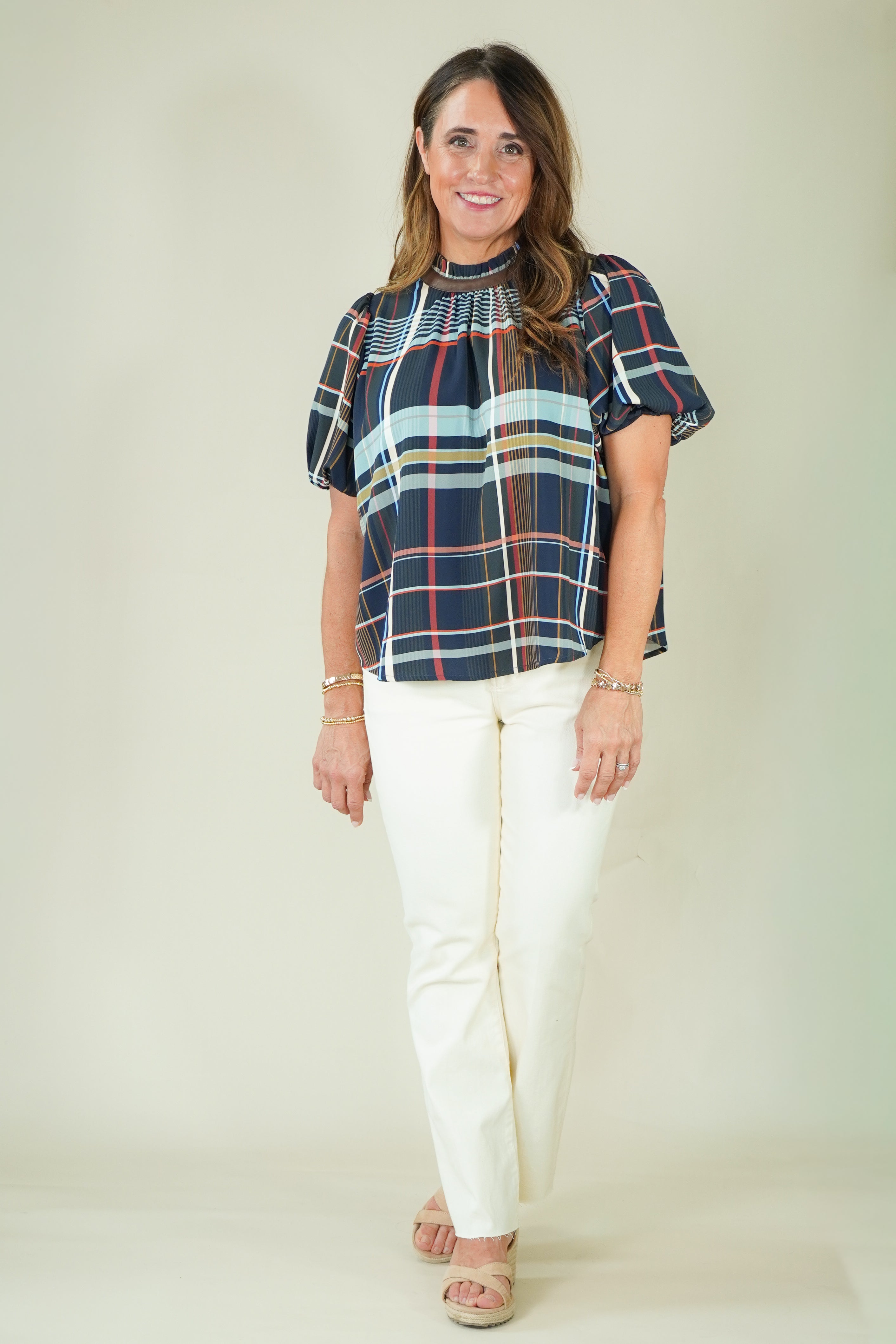 A woman modeling a short-sleeve plaid blouse with navy, red, and light blue stripes, paired with off-white pants and beige sandals, against a plain beige studio background.