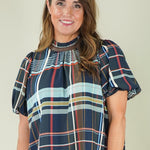 A woman modeling a short-sleeve plaid blouse with puff sleeves, featuring navy, red, and light blue stripes, paired with white pants and beige sandals, against a plain beige studio background.