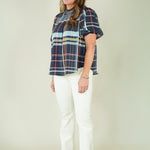 A woman modeling a short-sleeve plaid blouse with navy, red, and light blue stripes, paired with off-white pants and beige sandals, against a plain beige studio background.