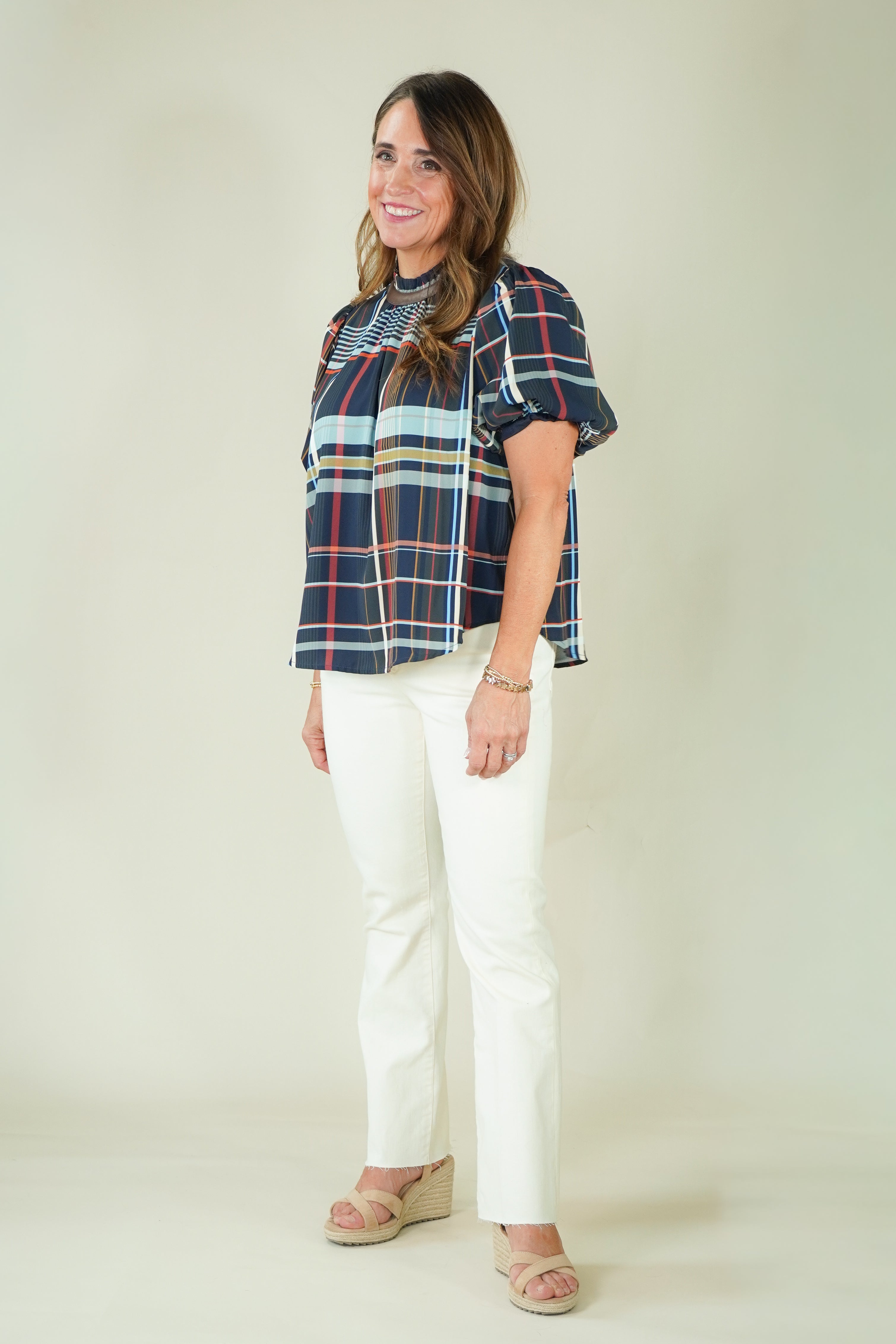 A woman modeling a short-sleeve plaid blouse with navy, red, and light blue stripes, paired with off-white pants and beige sandals, against a plain beige studio background.