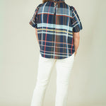 A woman modeling a short-sleeve plaid blouse with navy, red, and light blue stripes, paired with off-white pants and beige sandals, against a plain beige studio background.