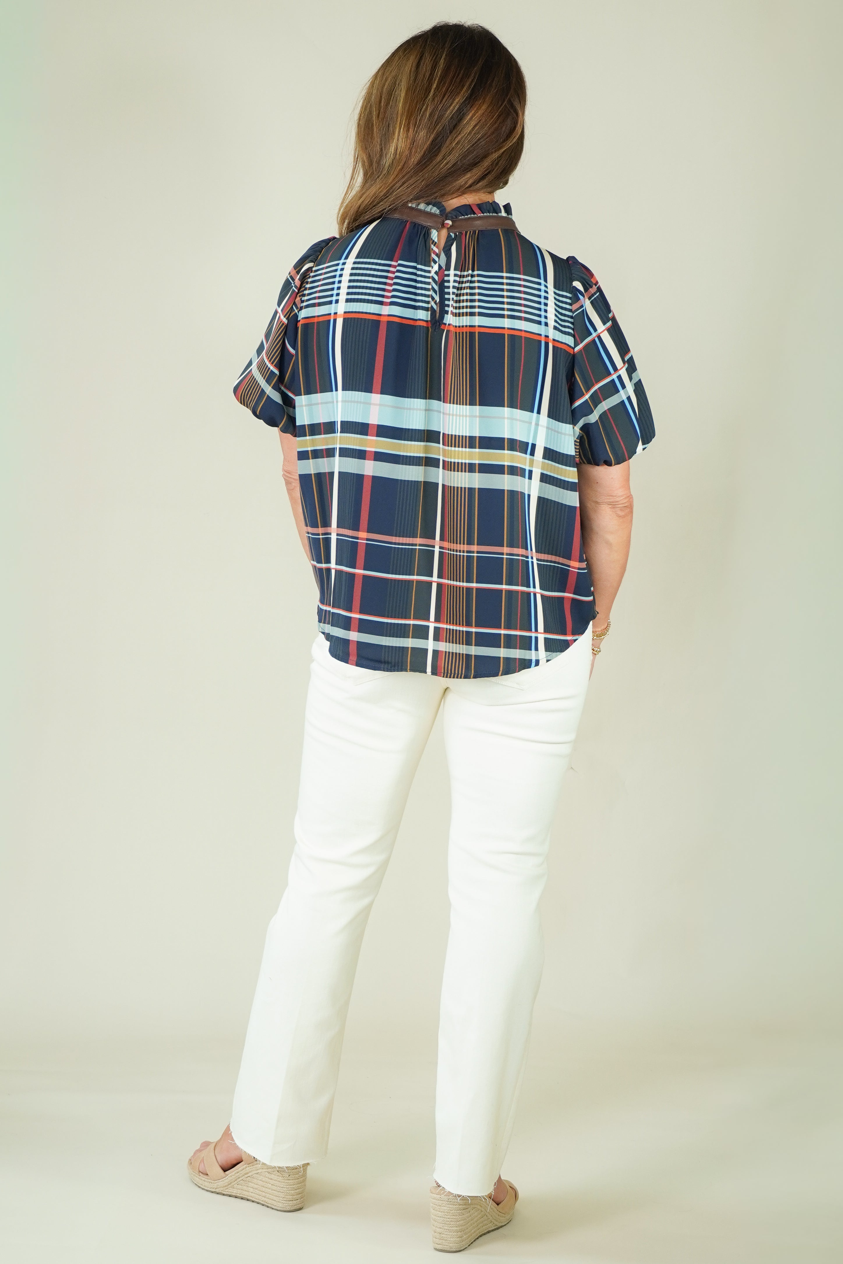 A woman modeling a short-sleeve plaid blouse with navy, red, and light blue stripes, paired with off-white pants and beige sandals, against a plain beige studio background.