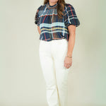 A woman modeling a short-sleeve plaid blouse with navy, red, and light blue stripes, paired with off-white pants and beige sandals, against a plain beige studio background.