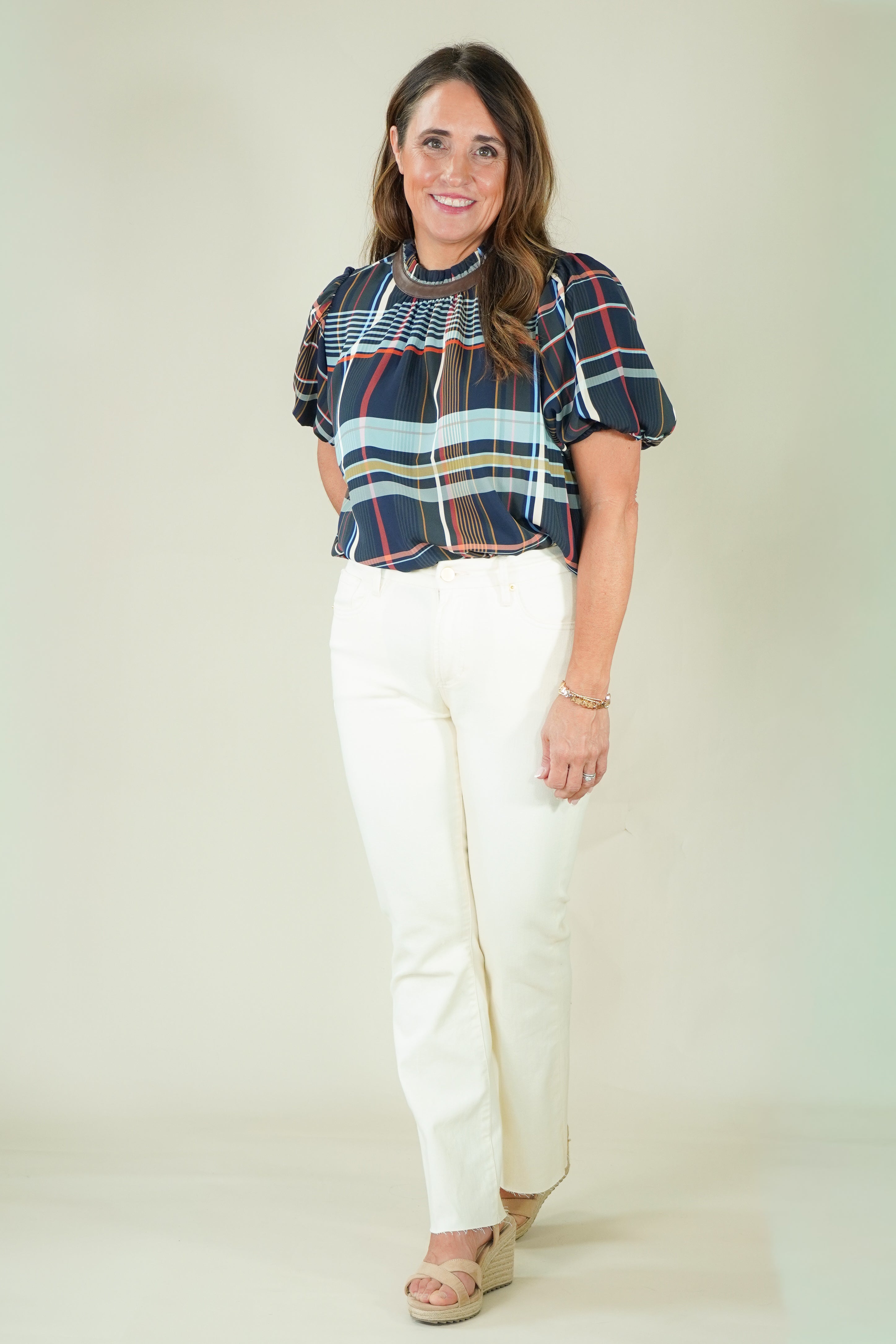 A woman modeling a short-sleeve plaid blouse with navy, red, and light blue stripes, paired with off-white pants and beige sandals, against a plain beige studio background.