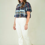 A woman modeling a short-sleeve plaid blouse with navy, red, and light blue stripes, paired with off-white pants and beige sandals, against a plain beige studio background.