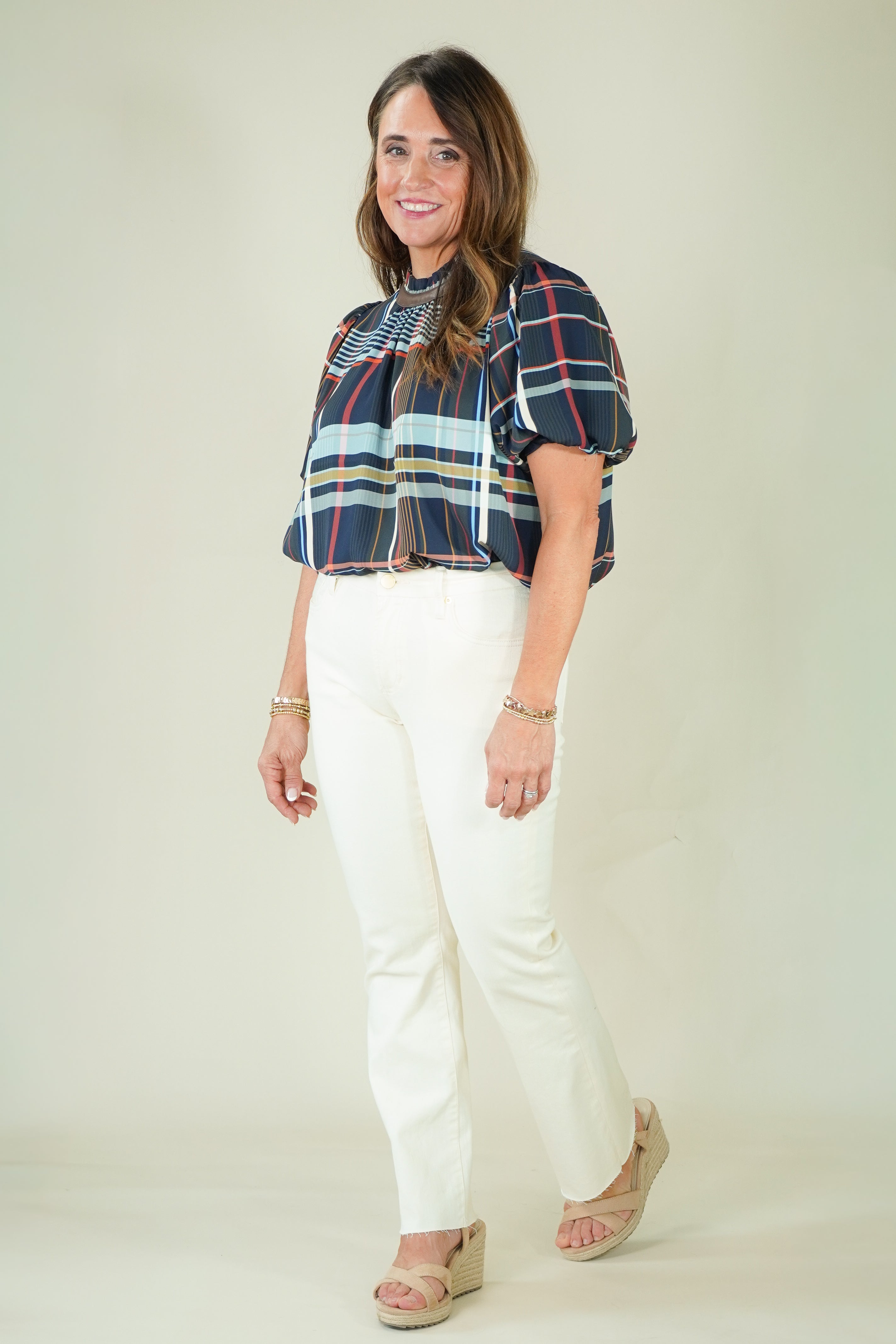 A woman modeling a short-sleeve plaid blouse with navy, red, and light blue stripes, paired with off-white pants and beige sandals, against a plain beige studio background.