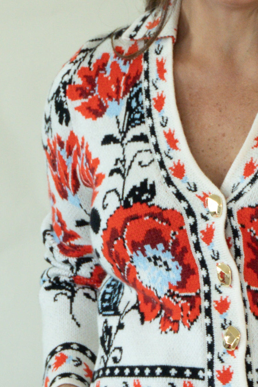 A woman modeling a white knit cardigan with vibrant red and black floral patterns, paired with dark blue jeans and tan boots, standing against a plain off-white studio background.