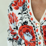 A woman modeling a white knit cardigan with vibrant red and black floral patterns, paired with dark blue jeans and tan boots, standing against a plain off-white studio background.